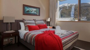 A hotel room with a bed, a nightstand, a lamp, a painting, and a window with a mountain view. - Squatter's Run Apartments (Thredbo)