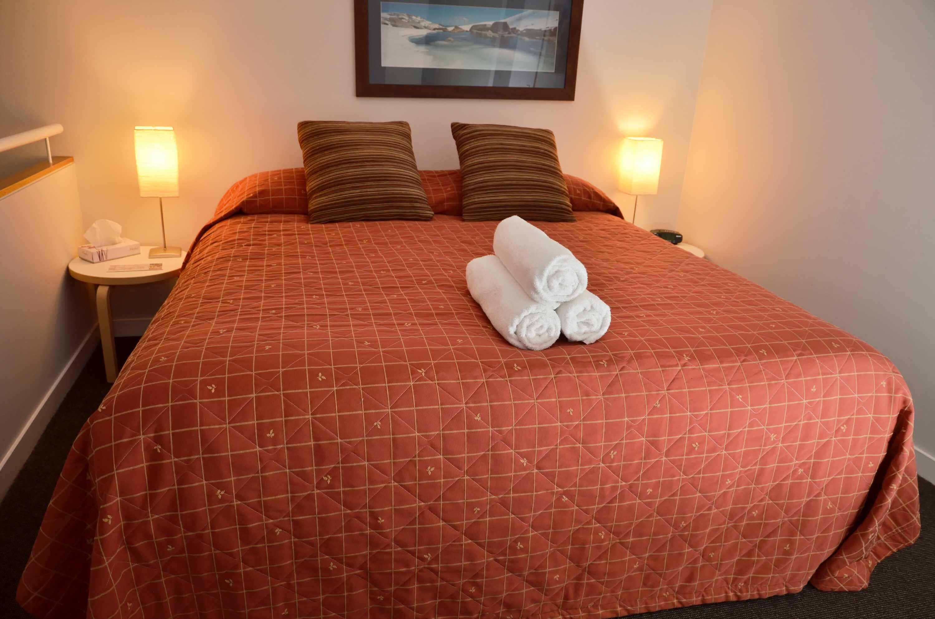A hotel room with a large bed, two pillows, a rolled towel, a bedside table with a lamp, and a framed picture on the wall.