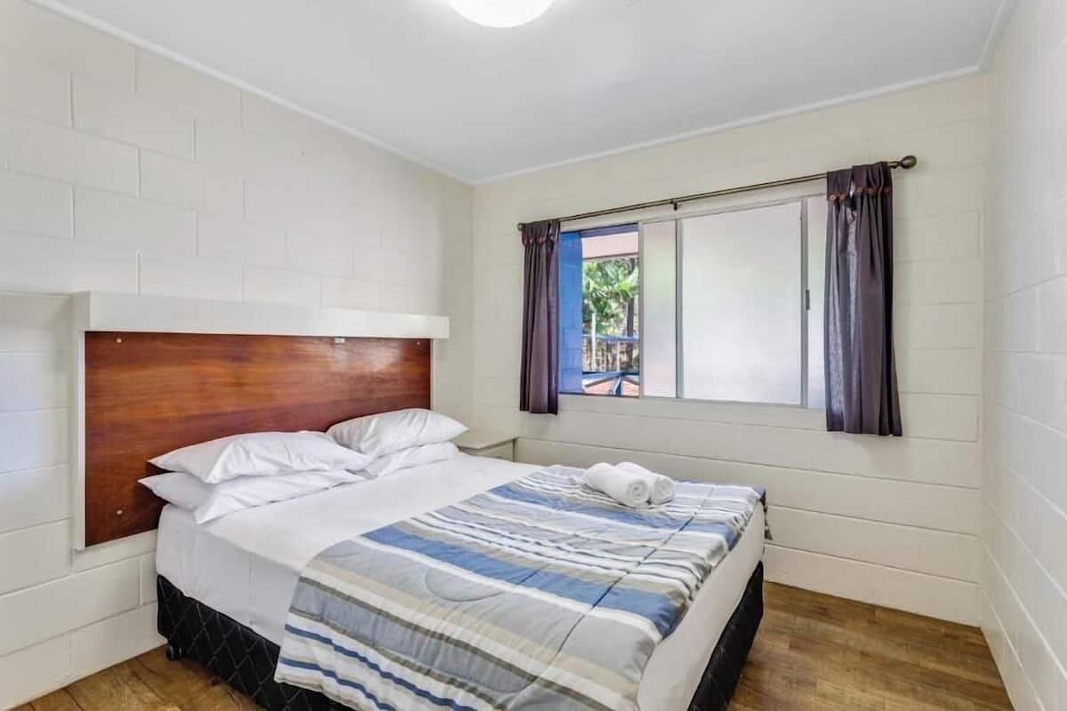 A bedroom with a wooden headboard, a bed with a striped bedspread, a window with curtains, and a view of the outside.