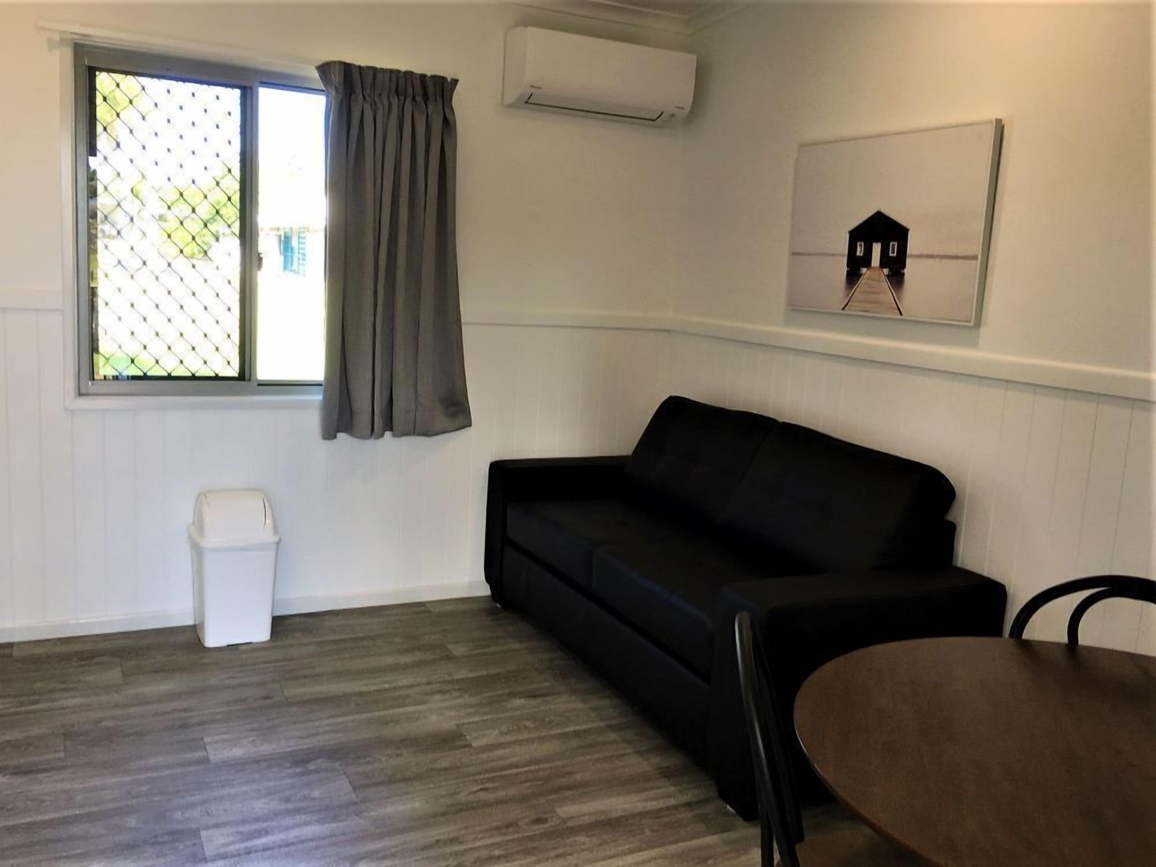 A room with a black sofa, a small white trash bin, a table, a window with a curtain, and an air conditioning unit on the wall.