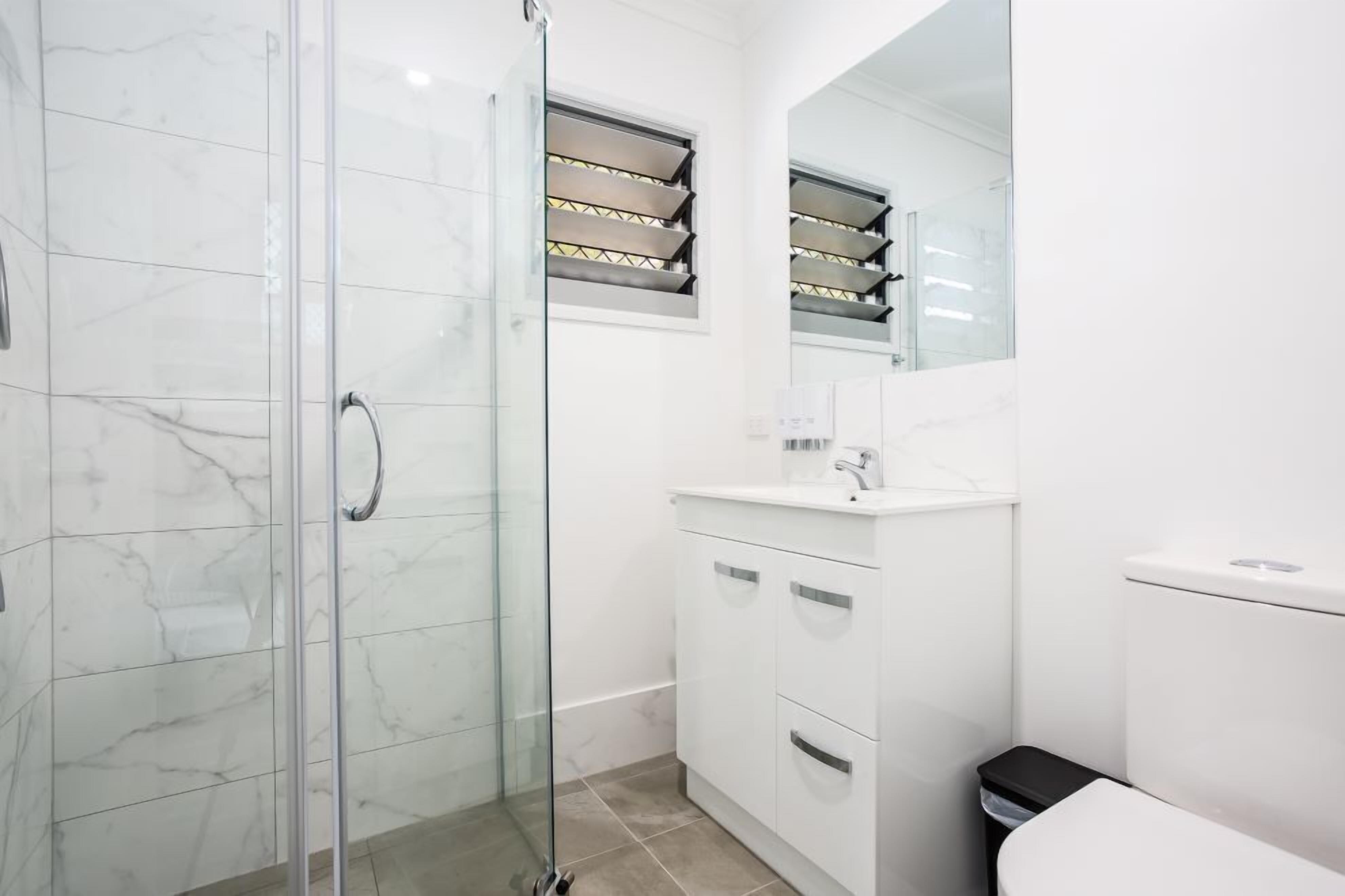 Een moderne badkamer met een glazen douchecabine, witte marmeren tegels, een wit wastafelmeubel en een zwarte prullenbak.