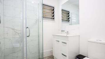 Een moderne badkamer met een glazen douchecabine, witte marmeren tegels, een wit wastafelmeubel en een zwarte prullenbak.