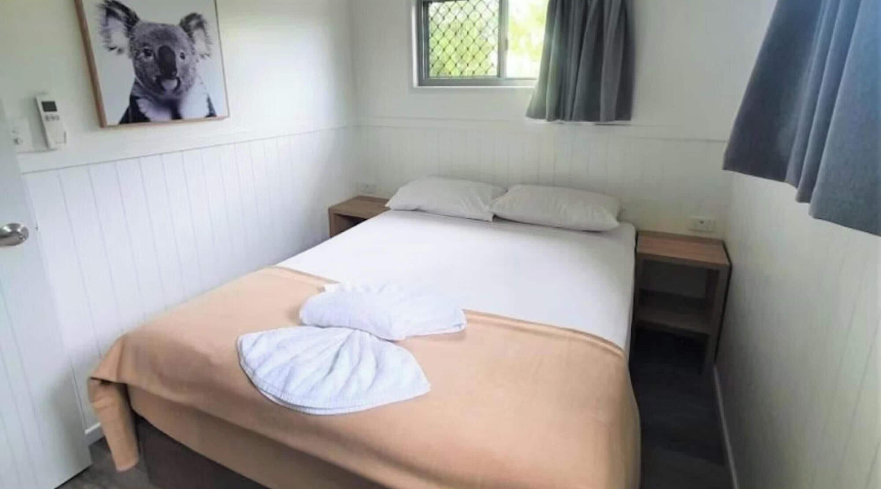 A single bed with white linens, a beige throw blanket, and a pillow. There is a window with a curtain, a wall-mounted thermostat, and a framed picture above the bed.