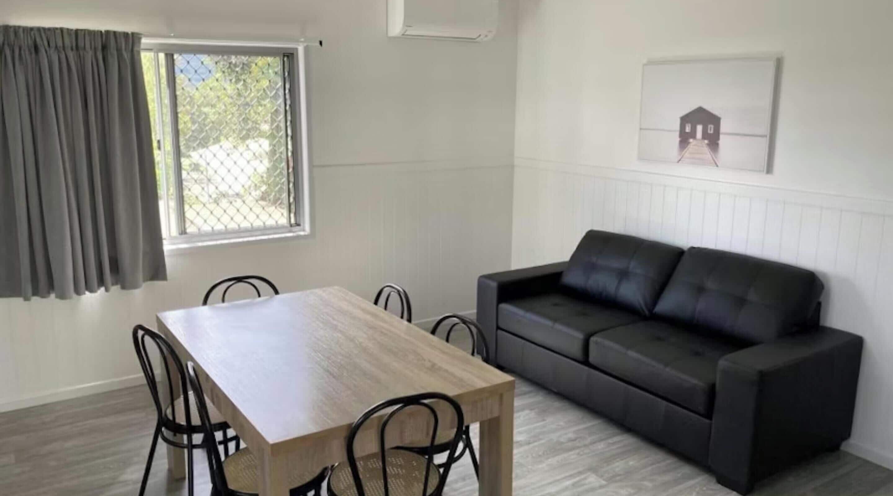 A room with a dining table, four chairs, a black sofa, a window with curtains, and a framed picture on the wall.