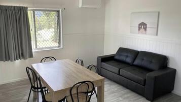 A room with a dining table, four chairs, a black sofa, a window with curtains, and a framed picture on the wall.
