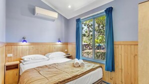 Une chambre à coucher avec une tête de lit en bois, un lit recouvert de draps blancs, une fenêtre avec des rideaux bleus et un climatiseur fixé au mur.