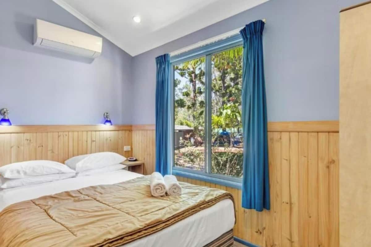 A bedroom with a wooden headboard, a bed with white linens, a window with blue curtains, and a wall-mounted air conditioner.