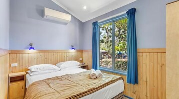 A bedroom with a wooden headboard, a bed with white linens, a window with blue curtains, and a wall-mounted air conditioner.