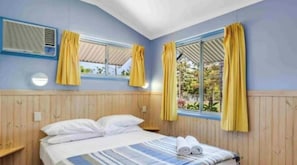 A hotel room with a bed, two windows with curtains, an air conditioning unit, and a view of greenery outside.