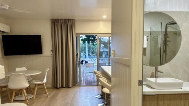 Une chambre d’hôtel moderne dotée d’une télévision à écran plat, d’un coin repas avec des chaises blanches, d’un bar avec des tabourets et d’un grand miroir rond au-dessus du lavabo.