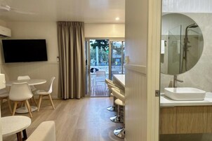A modern hotel room with a flat-screen TV, a dining area with white chairs, a bar area with stools, and a large round mirror above the sink.