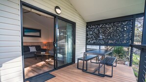 Una terraza de madera con puerta corrediza de vidrio que da a una sala de estar con un sofá, una mesita redonda y una lámpara.