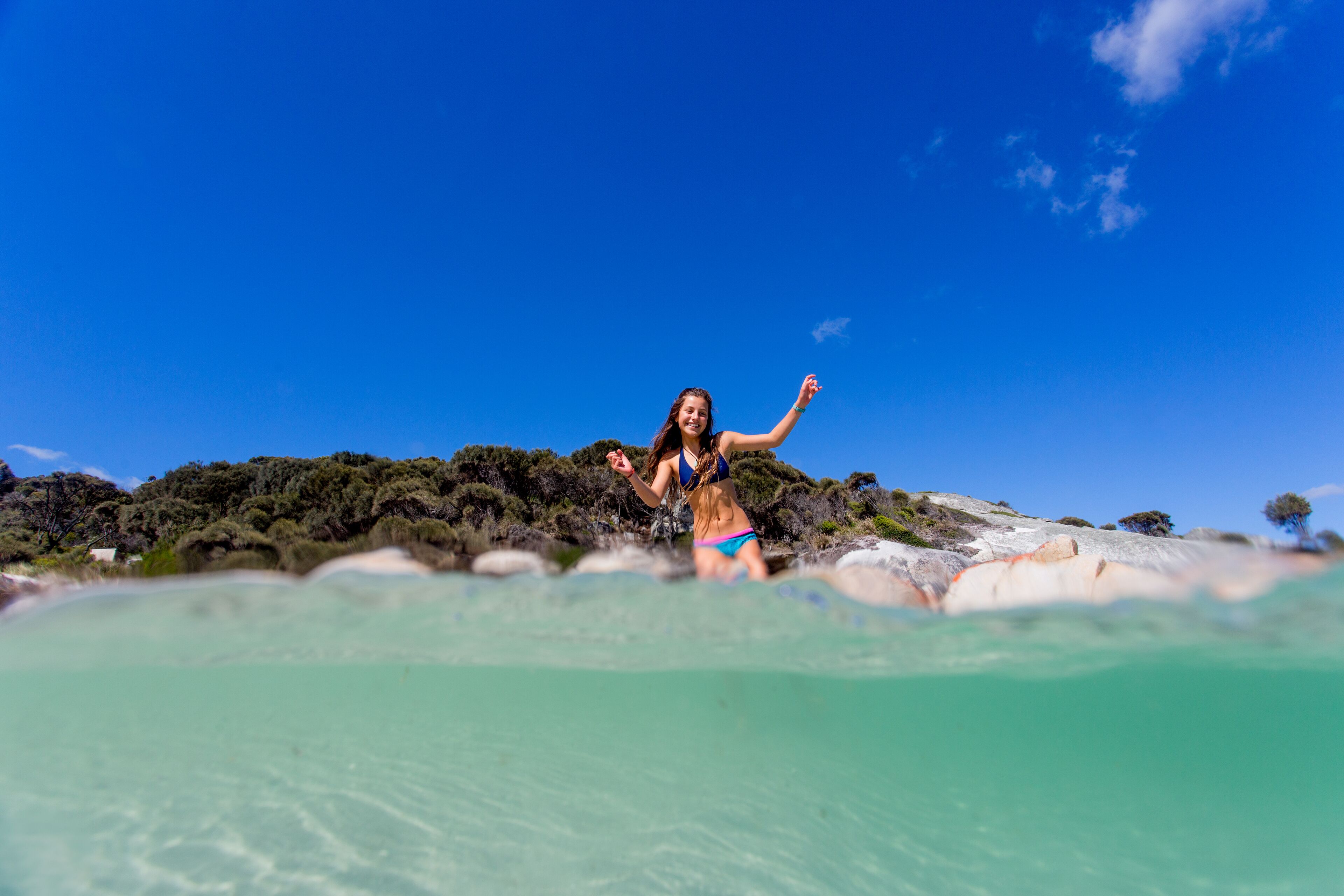 A person in a bikini standing on a rocky beach with clear turquoise water.