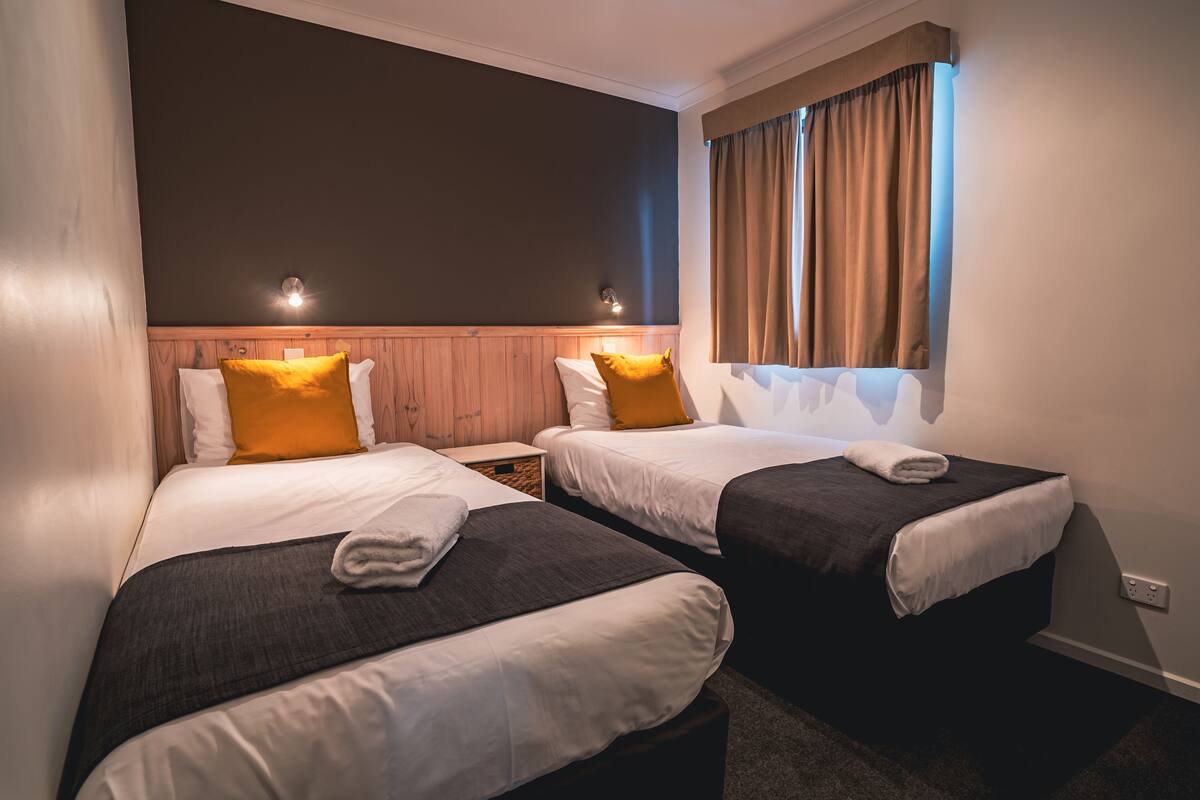 Quarto de hotel com duas camas, cada uma com travesseiro e cobertor. Há uma janela com cortinas e um ponto de luz fixado na parede.