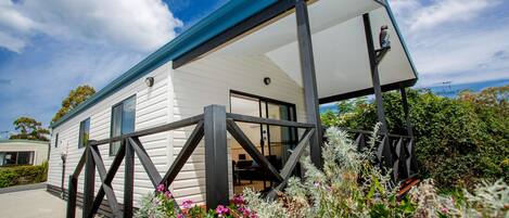 A white mobile home with a blue roof and a porch, surrounded by various plants and flowers.