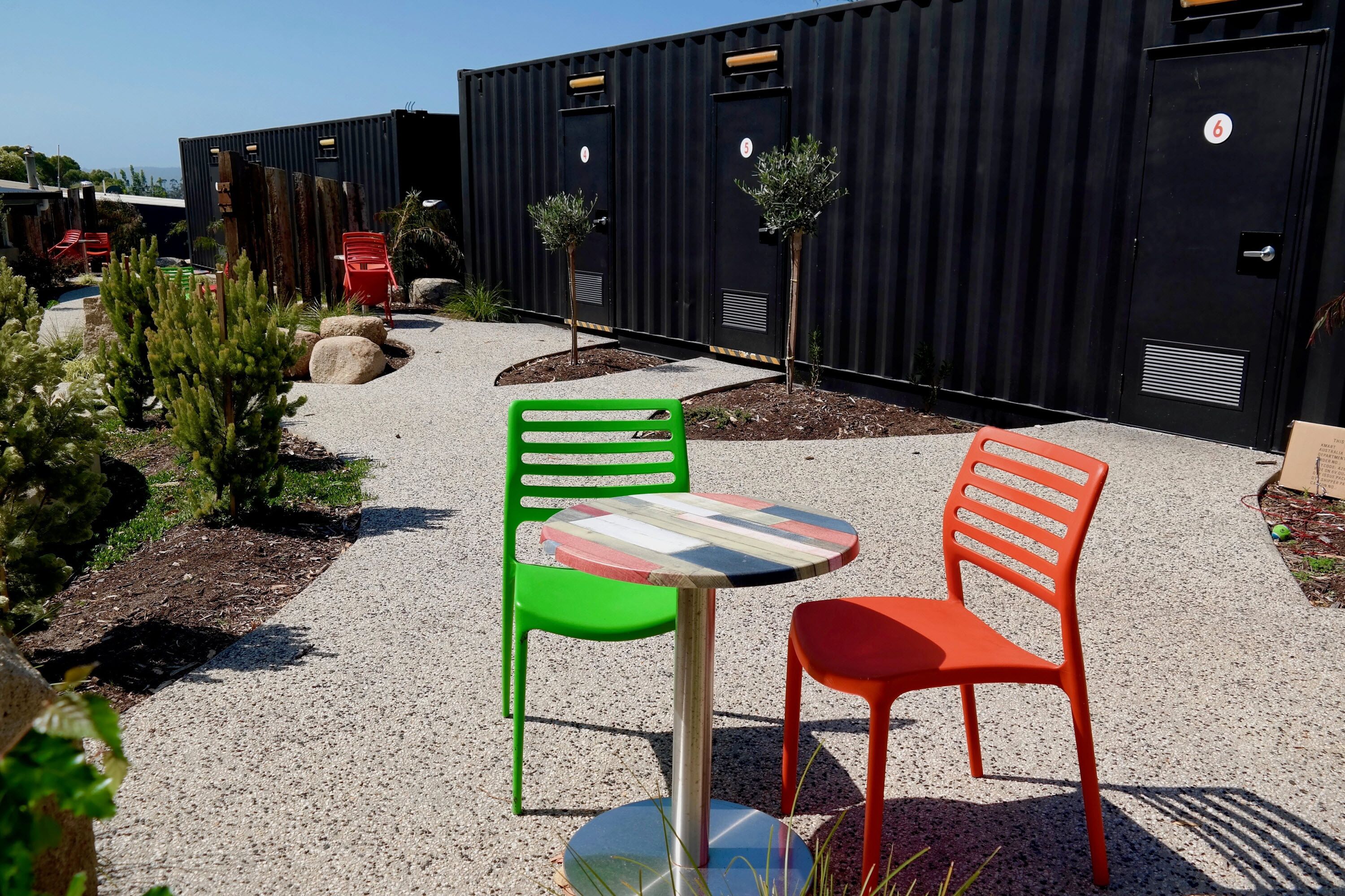 Une cour dotée d’un espace extérieur aménagé avec une table et deux chaises, entourée de verdure et bordée d’un bâtiment dont les portes sont numérotées.