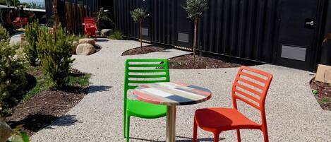 A courtyard with outdoor seating, including a table and two chairs, surrounded by greenery and a building with numbered doors.