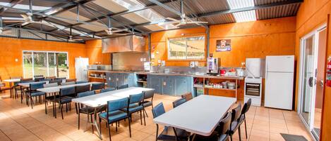 A spacious indoor dining area with high ceilings, multiple tables and chairs, a kitchen area with a refrigerator and stove, and a large window providing natural light.