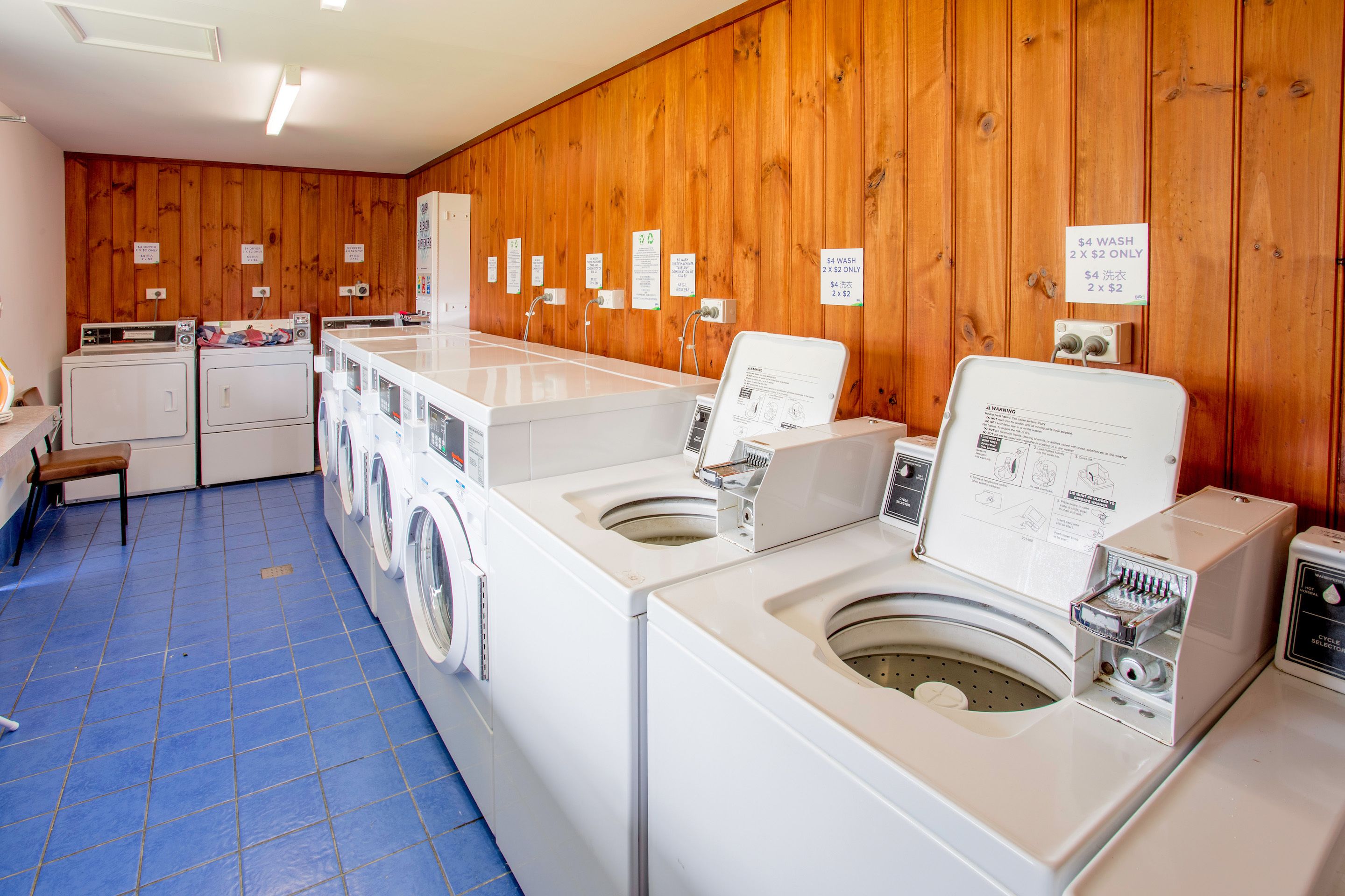 A laundry facility with multiple washing machines and dryers, wooden walls, and a blue-tiled floor.