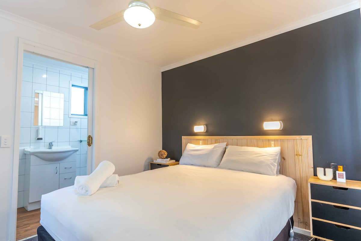 Quarto de hotel com cama, mesa de cabeceira, luminária na parede, ventoinha no teto, banheiro com pia e espelho, e uma porta.