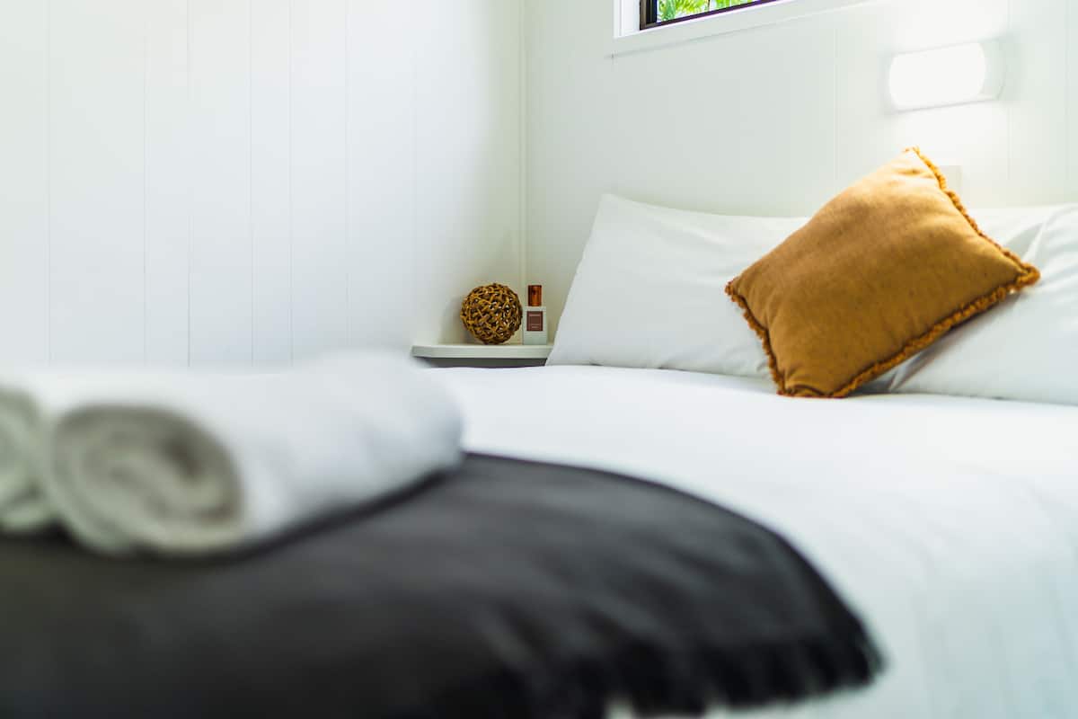 Cama bem arrumada com roupa de cama branca e preta, um travesseiro marrom e uma toalha branca enrolada em uma mesinha de cabeceira branca com um objeto decorativo.