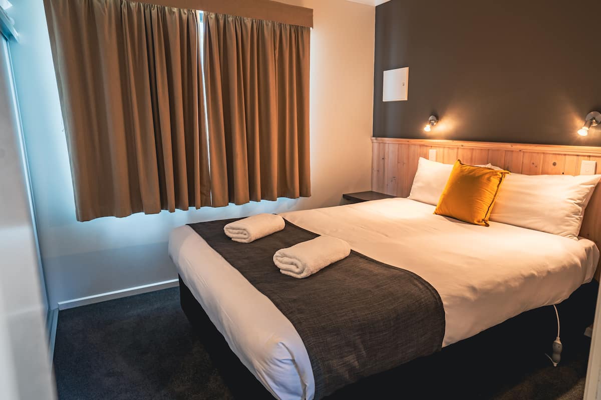 Quarto de hotel com uma cama grande, cabeceira de madeira, duas mesas de cabeceira e uma janela com cortinas.