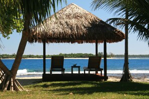 Un quiosco con techo de paja, con dos sillas de madera y una mesita, con vistas a una playa con palmeras y el océano.