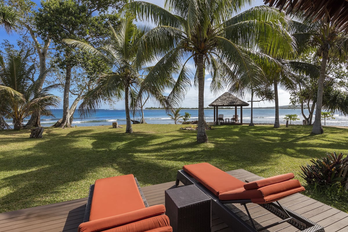 A deck with lounge chairs and a thatched-roof gazebo overlooking a beach with palm trees and the ocean.