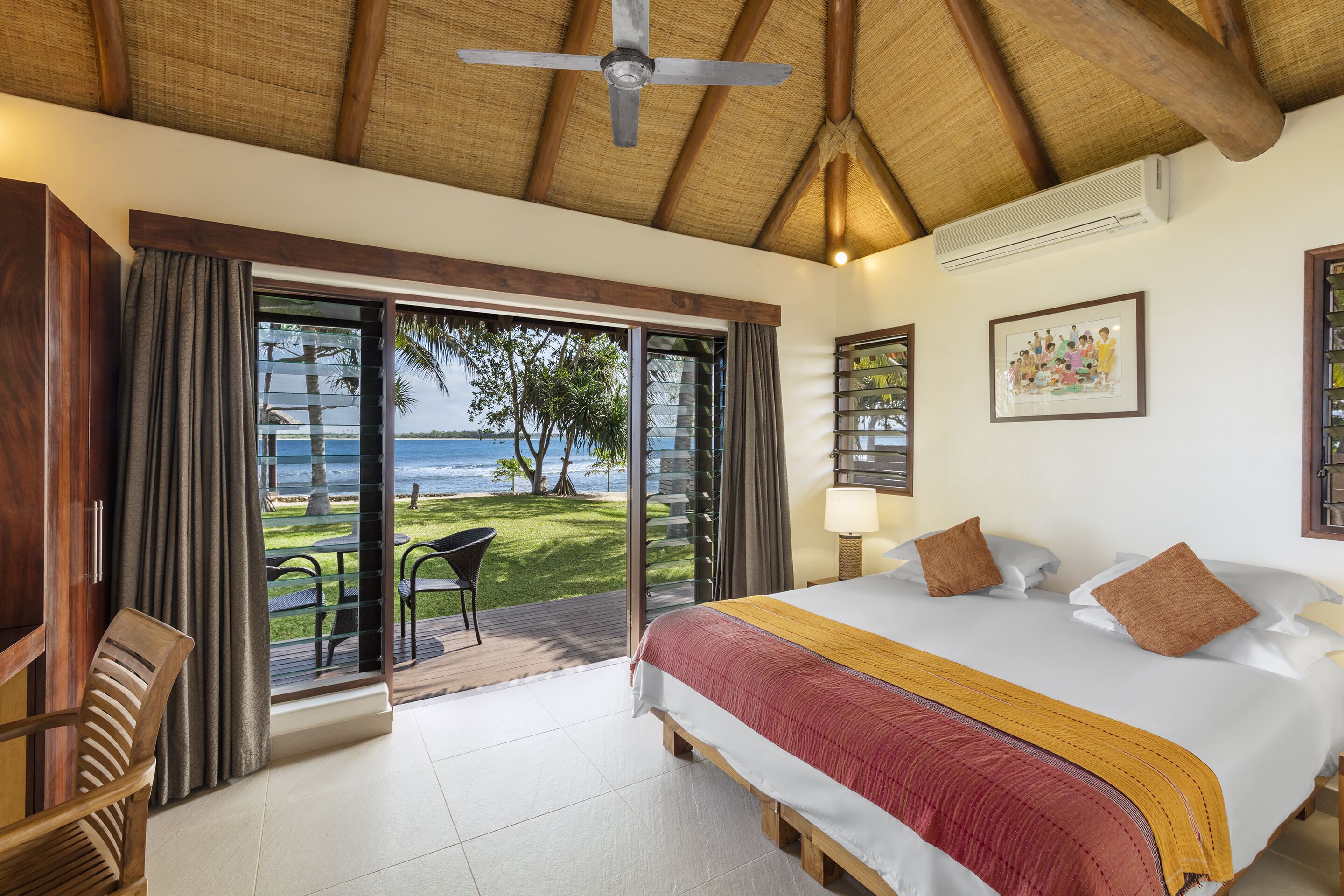 A bedroom with a large bed, a ceiling fan, a view of the ocean, and a balcony with outdoor seating.