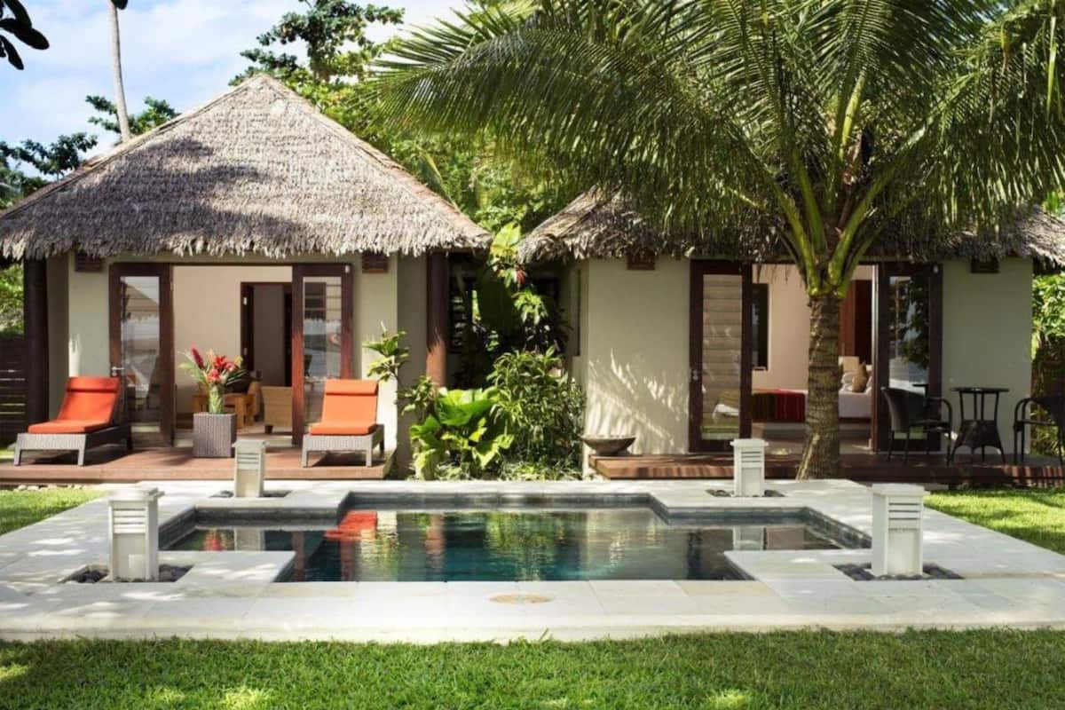 A pool area with two thatched-roof structures, each with a lounge chair and a small table.