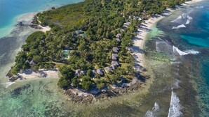 Vue aérienne d’un complexe hôtelier tropical situé sur une île, avec de nombreux hébergements de style bungalow entourés d’une végétation luxuriante, d’eaux turquoise cristallines et d’une plage de sable.