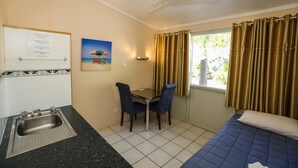 A compact room with a bed, a small desk with two chairs, a window with curtains, a sink, and a ceiling fan.