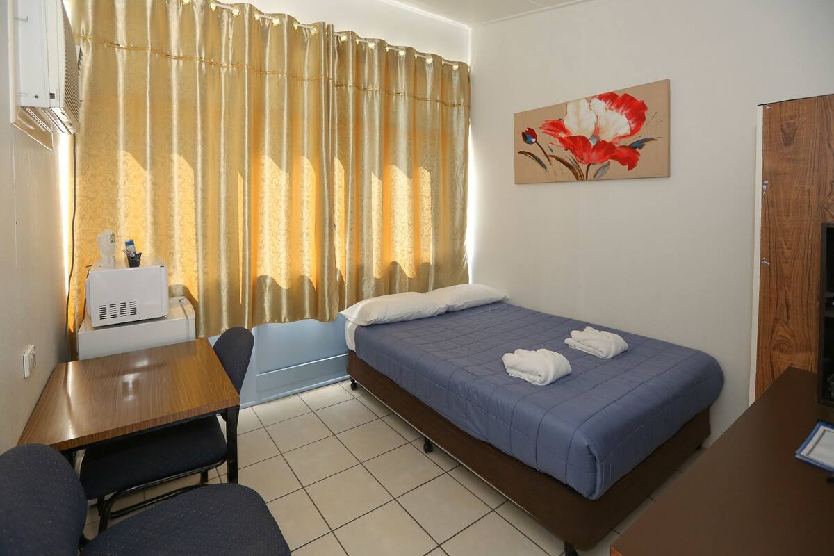 A hotel room with a bed, a desk, a chair, a small table, a microwave, and a painting on the wall.
