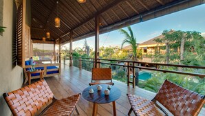 Uma área de estar coberta ao ar livre, com móveis de madeira e uma mesa, com vista para uma piscina e vegetação exuberante.