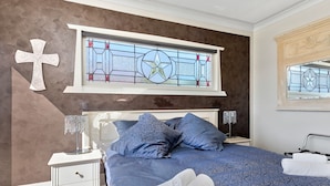 A bedroom with a large bed, two nightstands, a cross on the wall, and a stained glass window. - Lakeside City Apartments - Penthouse (Rotorua)