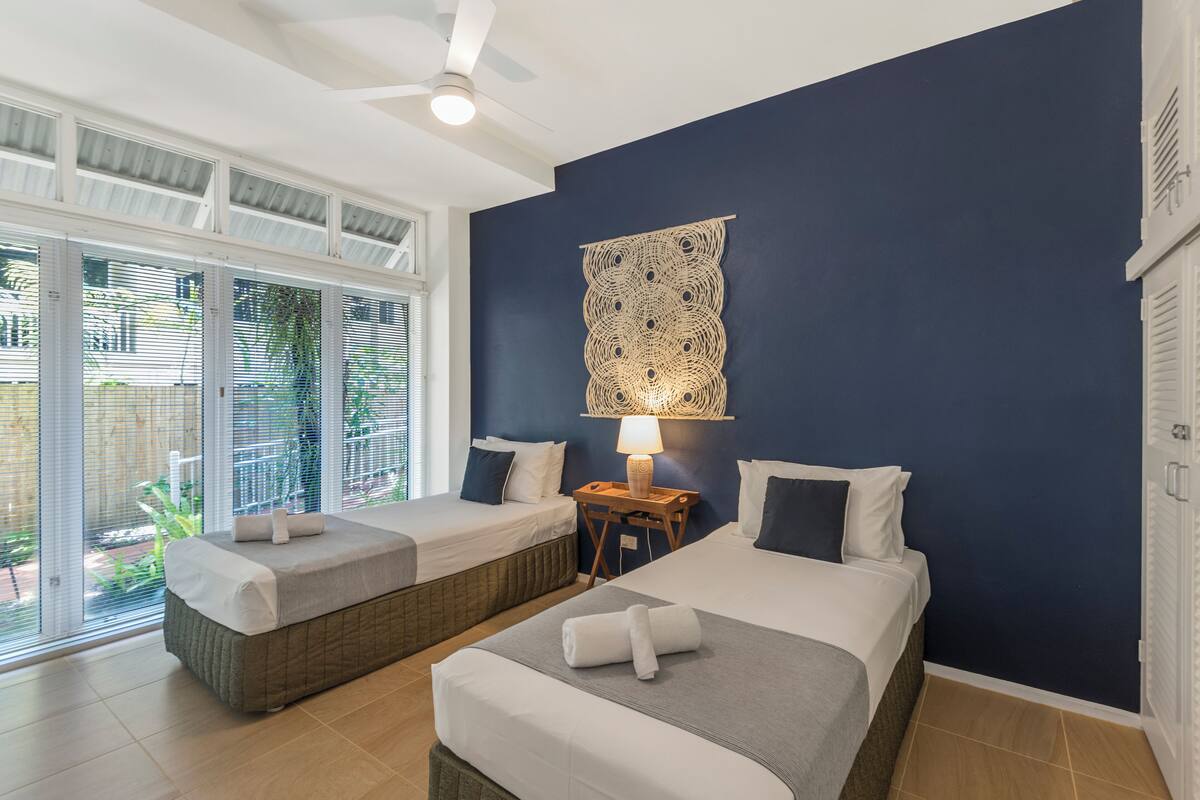 A hotel room with two beds, a ceiling fan, a large window with blinds, and a wall tapestry.