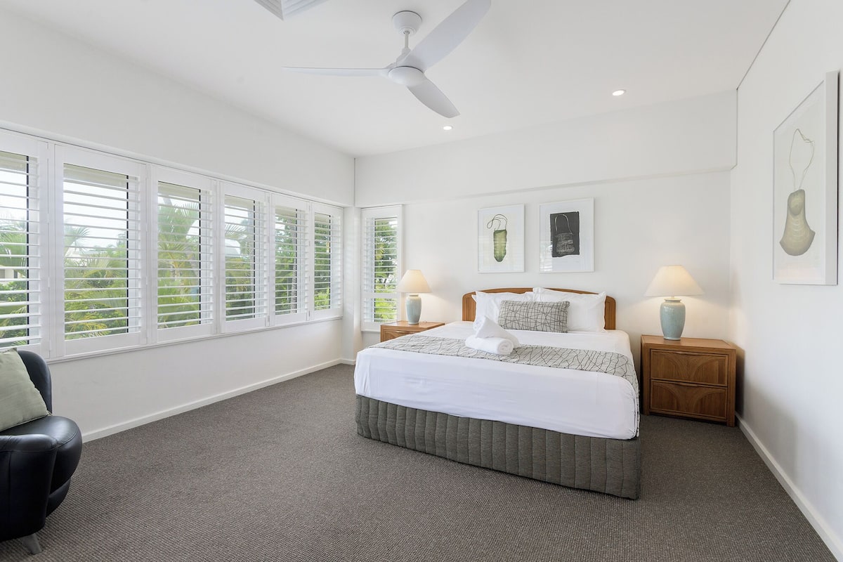 A bedroom with a bed, two bedside tables, a ceiling fan, and large windows with shutters.