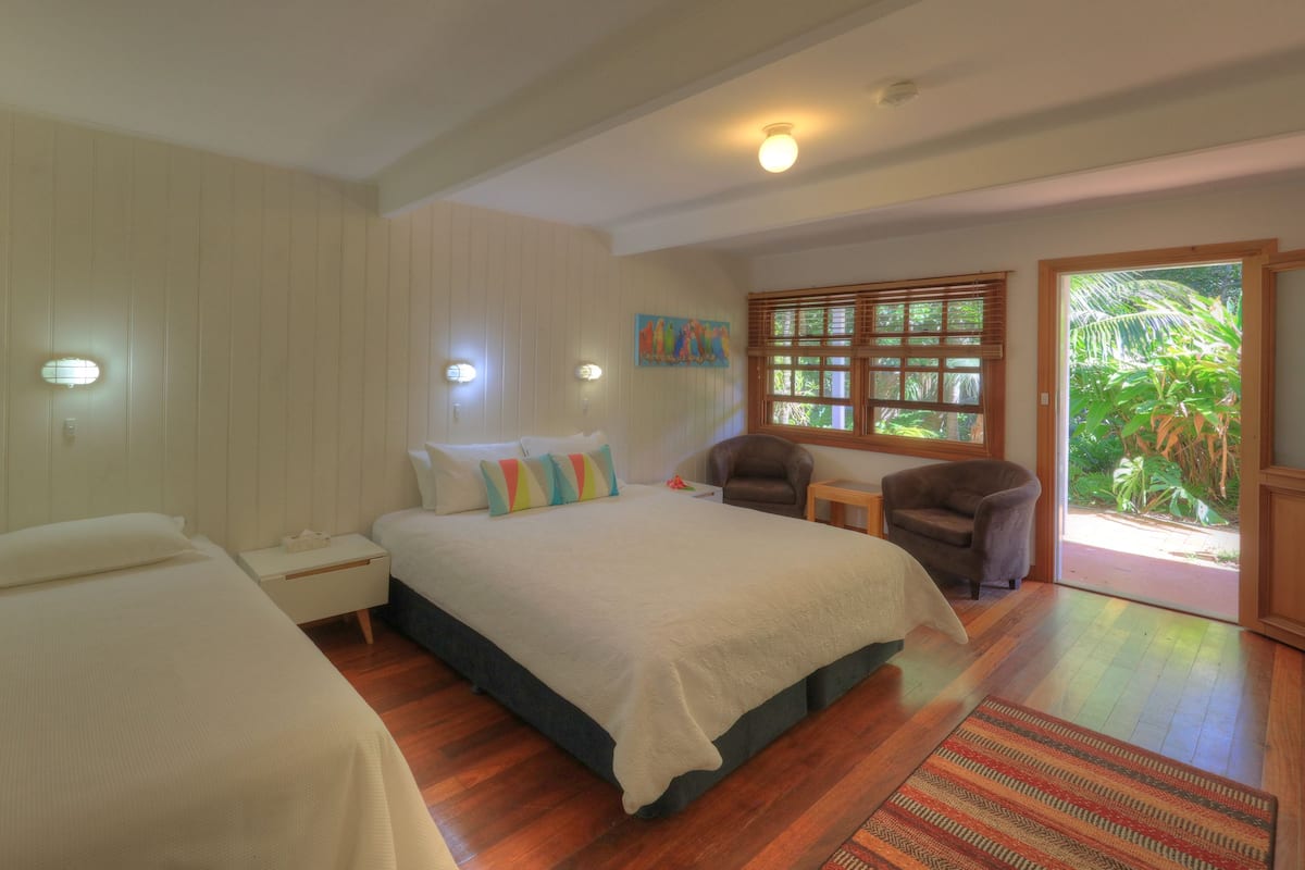 A bedroom with two beds, a small table, two armchairs, a window with blinds, and a door leading to a garden.