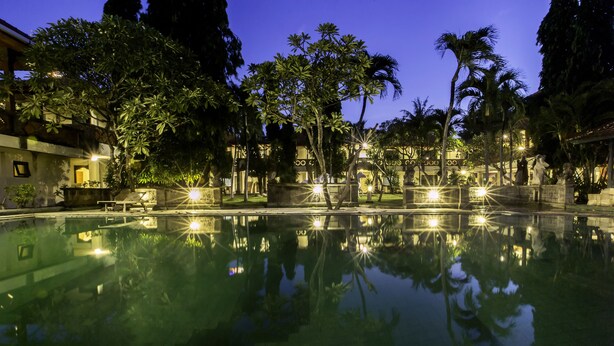 A nighttime pool area with reflections of palm trees and buildings, surrounded by well-lit pathways and a clear pool. - Bali Bungalo Hotel (Legian)