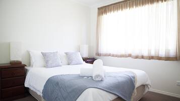 A neatly made bed with a rolled towel, a bedside table with a lamp, and a window with sheer curtains.