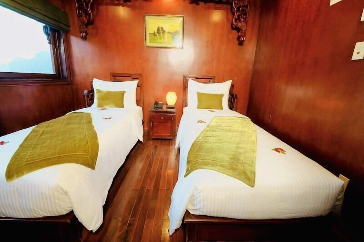A room with two beds, wooden flooring, a painting on the wall, and a nightstand with a lamp.