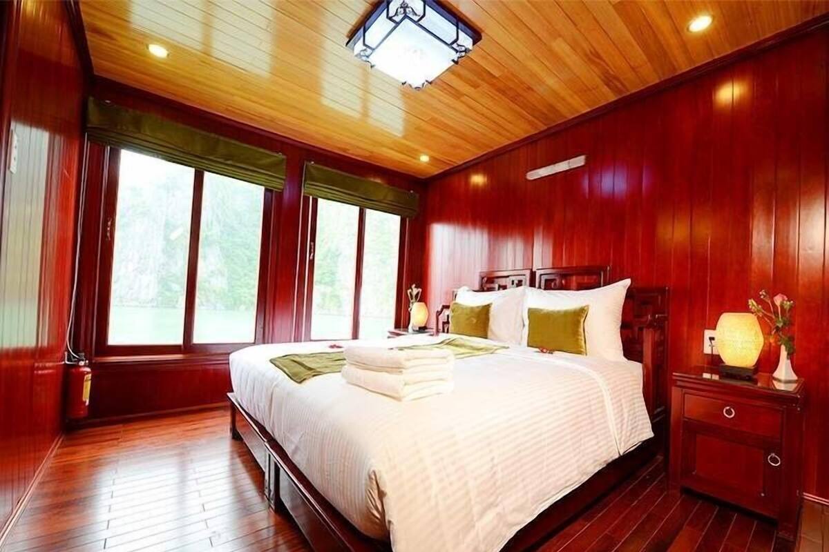 A spacious bedroom with a large bed, wooden flooring, and a view of the outdoors through large windows.