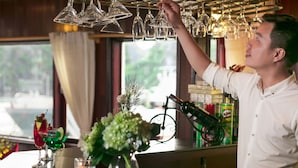 Una persona organizando copas de vino sobre una barra con vista a una ventana, un jarrón con flores y una botella de licor.