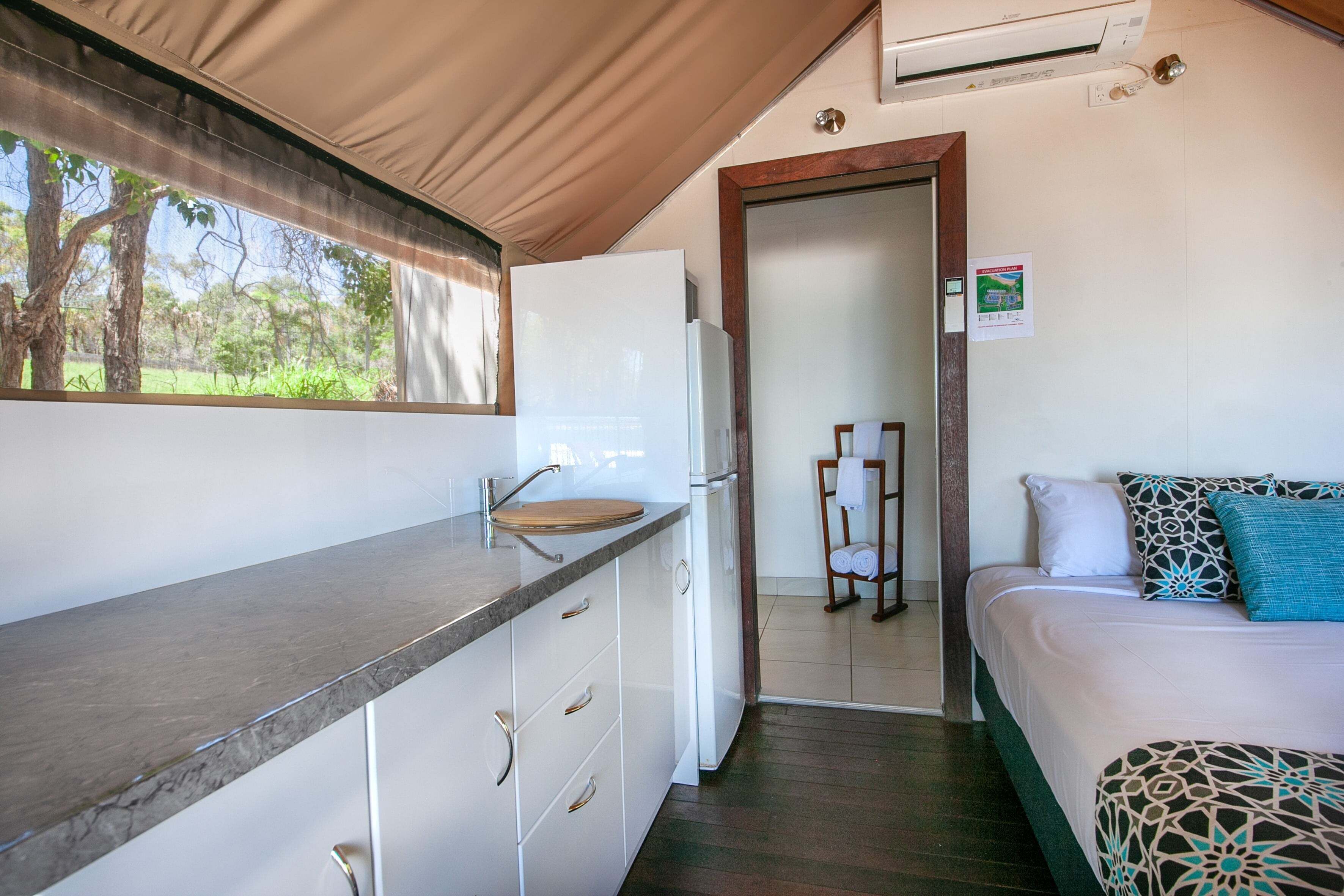 A compact kitchen with a sink, a refrigerator, and a wooden countertop. There is a bed with patterned bedding and a view of the outdoors through a window.