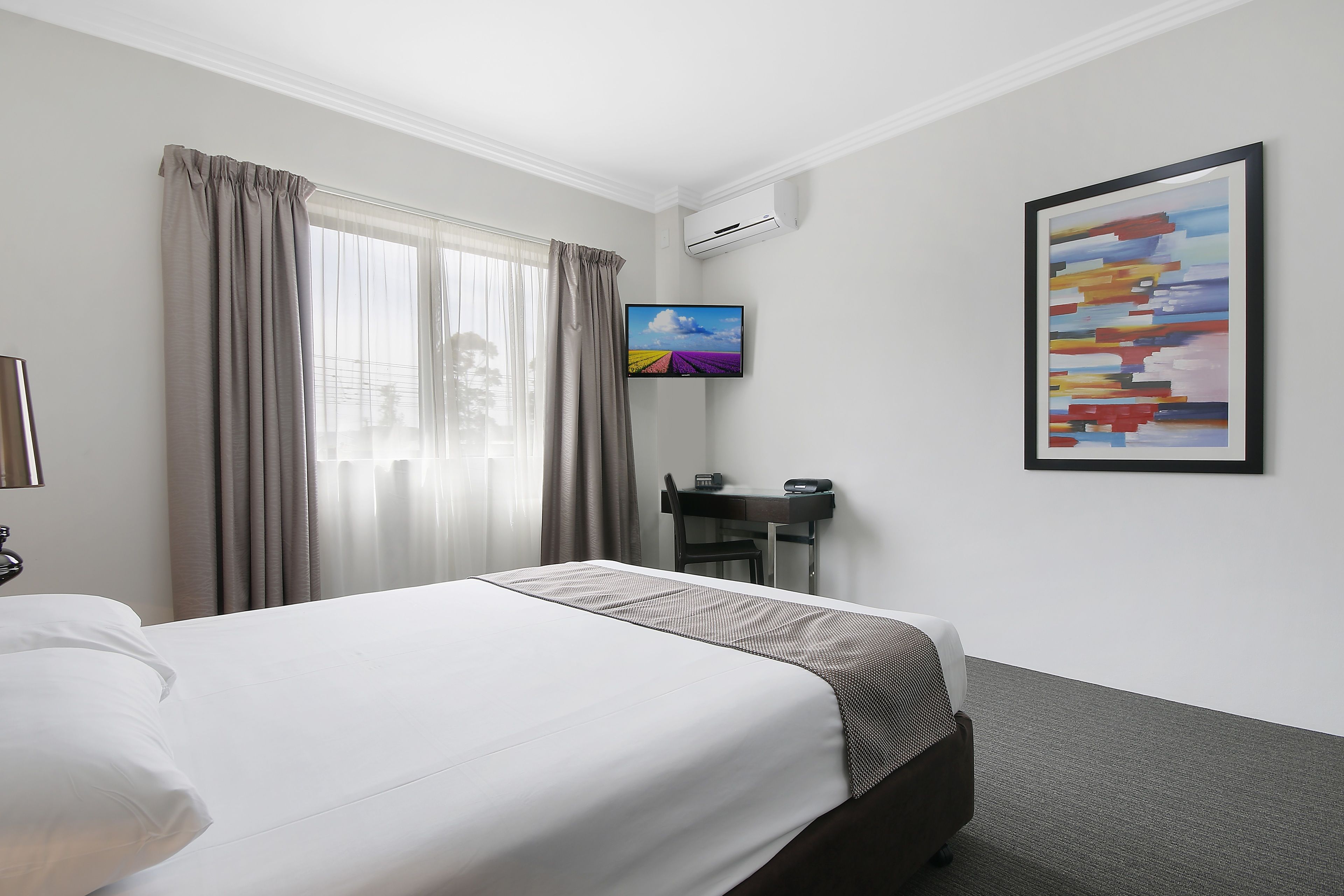 A hotel room with a bed, a desk, a chair, an air conditioner, and a painting on the wall.