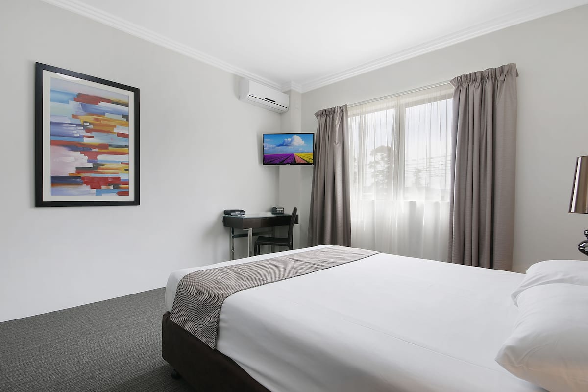 A hotel room with a bed, a desk, a chair, a TV, and a painting on the wall.