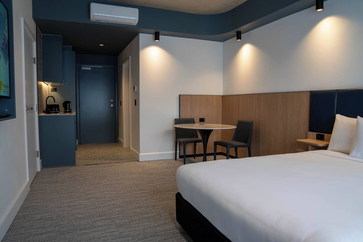 A modern hotel room with a large bed, a small round table with chairs, a kitchenette, and a separate door leading to a bathroom.