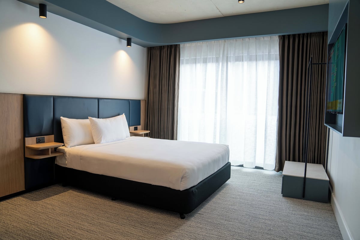 A modern hotel room with a large bed, bedside tables, a flat-screen TV, and a view of the outdoors through sheer curtains.