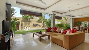 A spacious living area with a sofa, coffee table, and TV, a pool in the background, and a dining area with chairs and umbrellas.