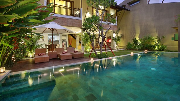 A poolside area with lounge chairs and umbrellas, a clear pool, and a building with balconies and outdoor lighting.